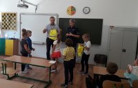 policjantka i pracownik WORD w Skierniewicach w szkole w klasie stoją przy tablicy, pokazują dzieciom odblaskowy workoplecak, przed policjantka i panem z WROD stoją dzieci
