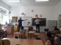 klasa szkolna, uczniowie siedzą w ławkach, policjantka oraz pracownik WORD w Skierniewicach są odwróceni tyłem do dzieci i mają wyprostowaną lewą rękę ucząc dzieci prawidłowo kierunków.