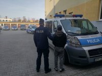 policjant prowadzi zatrzymanego do radiowozu, radiowóz stoi na parkingu dziedzińca komendy