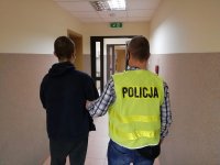 policjant w odblaskowej kamizelce konwojuje zatrzymanego mężczyznę do policyjnej celi, w tle korytarz i drzwi do pomieszczeń dla osób zatrzymanych