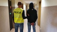 Policjant w kamizelce odblaskowej z napisem Policja prowadzi zatrzymanego. Korytarz w Kutnowskiej Komendzie Policji.