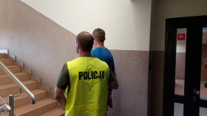 Policjant z odblaskową kamizelką z napisem Policja prowadzi po schodach zatrzymanego.