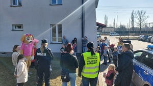 podwórko, po prawej stornie radiowóz, dzieci stoją na trawie, dwie pluszowe maskotki stoją wśród nich i policjantka