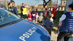 podwórko, po prawej stornie radiowóz, dzieci stoją na trawie, dwie pluszowe maskotki stoją wśród nich i policjantka