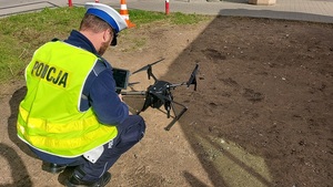 policjant kuca trzyma w ręku urządzenie sterujące drona na ziemi leży dron