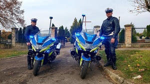 policjanci stoją obok motocykli za nimi brama wjazdowa na cmentarz