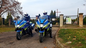 policjanci w kaskach siedzą na motocyklach, za nimi brama wjazdowa na cmentarz