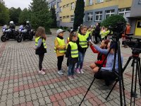 dzieci udzielają wywiadu do telewizji, z boku widać kamery na statywach, w tle pozostałe dzieci z klasy ora motocykle policyjne