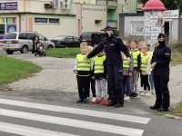 policjantka stoi z dziecmi w kamizelkach odblaskowych przed przejściem dla pieszych