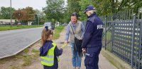dziewczynka wręcza opaskę odblaskową kobiecie na ulicy, obok stoi policjantka