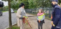 dziewczynka wręcza opaskę odblaskową kobiecie na ulicy, obok stoi policjantka