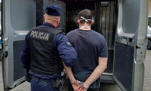 policjant stoi przed radiowozem oznakowanym trzyma zatrzymanego, który ma założone kajdanki na ręce z tyłu
