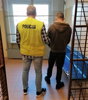 korytarz komendy policji, policjant w żółtej kamizelce odblaskowej prowadzi zatrzymanego mężczyznę