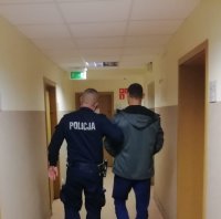 policjant w granatowym mundurze  z  napisem na plecach policja prowadzi korytarzem komendy zatrzymanego mężczyznę