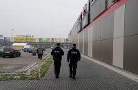 policjanci idą wzdłuż pasażu handlowego, po lewej stronie widać parking samochodowy, liczne samochody zaparkowane