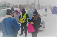 Policjant w kamizelce odblaskowej i redektorka Radia Q na Placu Piłsudskiego  rozdają odblaski mieszkańcom miasta, w tle radiowóz policyjny i choinka, kolejka mieszkańców.