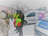 Policjant w kamizelce odblaskowej i redektorka Radia Q na Placu Piłsudskiego  rozdają odblaski mieszkańcom miasta, z prawej strony widać radiowóz policyjny oraz samochód radia Q