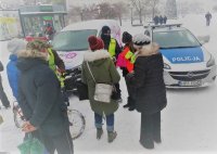 Policjant w kamizelce odblaskowej i redektorka Radia Q na Placu Piłsudskiego  rozdają odblaski mieszkańcom miasta, w tle radiowóz policyjny i samochód radia Q, kolejka mieszkańców.