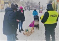 Ulica Królewska, mama ciągnie dziecko na sankach, policjant o redaktorka Radia Q przekazują dziecku odblaski.