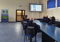 na sali odpraw siedzą za stołami policjanci, na ekranie multimedialnym na ścianie wyświetlana jest prezentacja