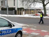 policjant z Ruchu Drogowego stoi na drodze, prawą rekę ma uniesioną ku górze , w ręku trzyma lizaka sygnalizując aby pojad zatrzyamał się.  Na pierwszym planie widać kawałek radiowozu policyjnego