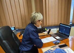 policjantka siedzi w pokoju za biurkiem, prowadzi zajęcia on line z młodzieżą, na biurku stoi laptop ono kalendarz, na biurku widać różne dokumenty.