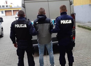 dwaj policjanci prowadzą na dziedzińcu komendy zatrzymanego do radiowozu