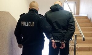 Policjant umundurowany prowadzi na klatce schodowej zatrzymanego. Zatrzymany ma założone kajdanki na ręce trzymane z tyłu.