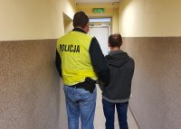 policjant w kamizelce odblaskowej stoi na korytarzu komendy trzymając niższego do niego mężczyznę-zatrzymanego