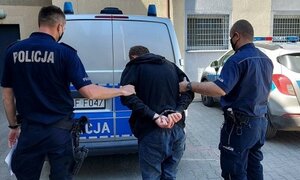 dziedziniec Komendy Powiatowej Policji w Kutnie, dwaj policjanci prowadzą do radiowozu zatrzymanego. Zatrzymany ma założone kajdanki na ręce trzymane z tyłu. Przed policjantami i zatrzymanym stoi radiowóz