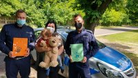 radiowóz policyjny, przed radiowozem stoi dwóch policjantów w rękach trzymają pamiątkowe laurki. Pomiędzy policjantami stoi pani i trzyma na reku synka oraz pluszowego misia