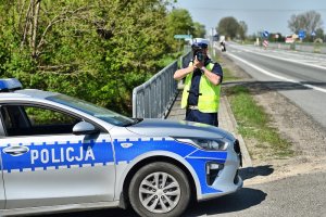 policjant stoi na drodze i mierzy prędkość urządzeniem obok widać radiowóz policyjny