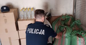 garaż, policjant kuca tyłem w umundurowaniu służbowym, przed nimi leżą pudełka kartonowe na nich butelki plastikowe z białą cieczą, po prawej stronie worki wypełnione krajanką tytoniową.