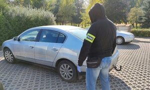 na parkingu stoi srebrny vw passat, obok stoi tyłem policjant z kapturem na głowie na ręce opaska z napisem Policja