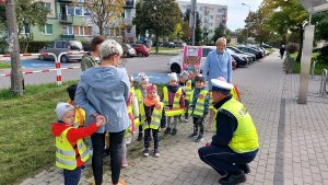 chodnik, po chodniku idzie grupa przedszkolaków w kamizelkach odblaskowych, policjant kuca przy nich rozmawia.