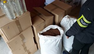 garaż pudełka kartonowe na wierzchu plastikowe butelki z bezbarwna cieczą na podłodze stoi worek jutowy wypełniony  tytoniem obok policjantka z opaską odblaskową z napisem Policja