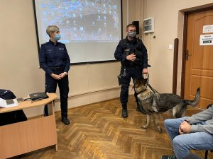 policjant i policjantka stoją w klasie przy tablicy prowadza prelekcje jest obok również pies służbowy, młodzież siedzi na krzesłach