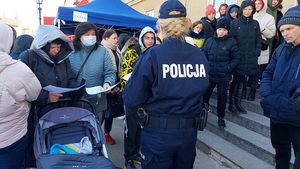 policjanta stoi przed urzędem miasta i rozdaje uchodźcom ulotki , przed budynkiem tłum ludzi.