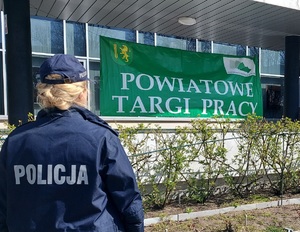 baner zielony Powiatowe Targi Pracy przed nim tyłem stoi policjantka