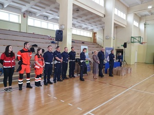 na sali gimnastycznej stoją ratownicy medyczni policjanci w oddali widać poukładane nagrody i puchary