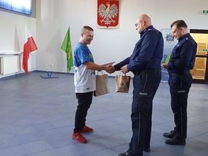 Komendant Powiatowy Policji w Kutnie wręcza upominek mieszkańcowi, ściska mu dłoń, za komendantem stoi policjant.