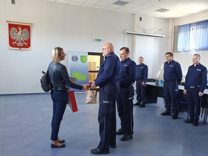 Komendant Powiatowy Policji w Kutnie wręcza upominek mieszkance, ściska jej dłoń, za komendantem stoi policjant.
