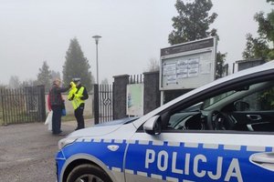 widać kawałek radiowozu, policjanta w bramie cmentarza rozdaje kamizelki odblaskowe