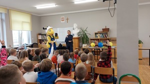 klasa dzieci siedzą na krzesłach z przodu pani policjantka i pluszowa maskotka mrówka