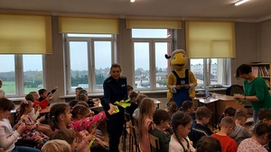 dzieci siedzą na krzesłach, policjantka rozdaje odblaski, z daleka stoi pluszowa maskotka mrówka