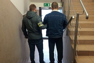 Policjant prowadzi po schodach w dół zatrzymanego. Po prawej tronie schody w górę.