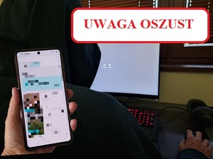 osoba z zaciągniętym kapturem siedzi przed monitorem komputerowym. Na pierwszym planie dłoń trzymająca telefon.