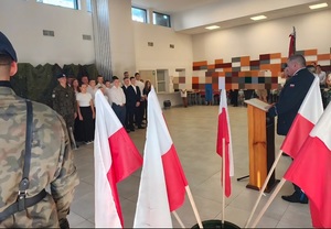 Komendant Powiatowy Policji w kutnie podczas przemówienia. W tle flagi Polski oraz uczniowie z klasy mundurowej.