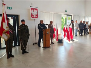 Komendant Powiatowy Policji w kutnie podczas przemówienia. W tle flagi Polski oraz uczniowie z klasy mundurowej.