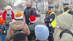 Policjanci z Mikołajem przekazują grupie dzieci elementy odblaskowe.